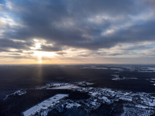 Winter aerial epic view on snowy village in forest. Zmiyevsky region in Ukraine from drone. Sun shines through heavy sunset clouds