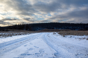Fototapeta na wymiar Snowy white rural road in countryside. Evening winter landscape with epic heavy clouds sky, reeds and dark trees forest hill