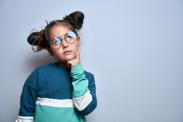 Fototapeta Funny little girl in glasses holding chin doubts, makes decision isolated on yellow studio background obraz