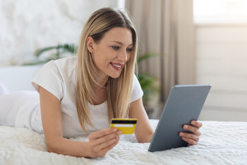 Positive woman laying in bed with tablet and credit card
