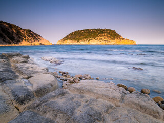Fototapeta na wymiar Paisaje costero con rocas en primer plano y una isla de fondo sobre cielo azul al atardecer