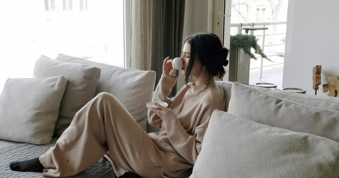 A beautiful Young woman is drinking coffee and tea in the living room at home, healthy calm lady relaxing on comfortable sofa feel stress free at home lounge alone. Woman rests at home on luxury sofa.