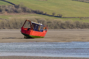A rusting fishing trawler lays stranded on a sandbank in a tidal estuary. River Teifi, with hilly farmland background. Rippling blue water. Landscape image with space for copy. Poppit Sands, Wales. - 502026423
