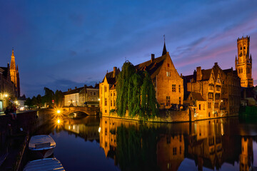 Fototapeta premium Famous view of Bruges tourist landmark attraction - Rozenhoedkaai canal with Belfry and old houses along canal with tree in the night. Brugge, Belgium