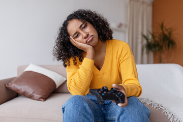 Unhappy young African American woman with joystick losing video game, sitting on sofa at home