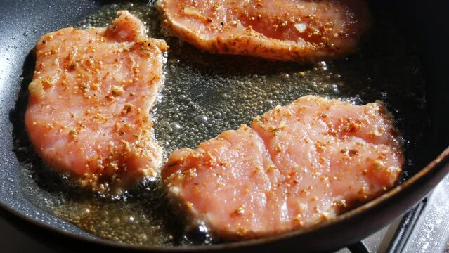 A large piece of pork schnitzel meat with seasonings is fried in a pan in a home kitchen. Close up view