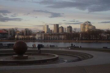 Obraz premium Full-color horizontal photo. Evening landscape in the city on the river bank. Sunny weather. Winter has just ended. Trees stand with bare branches. Massive concrete structures and large city buildings