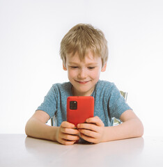 the boy holds a smartphone in his hands and enthusiastically looks at its screen. sitting at a table, white background.