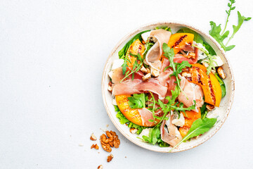 Summer salad bowl with sweet grilled peach, jamon, soft cheese, walnuts and fresh arugula on white kitchen table background, top view, negative space