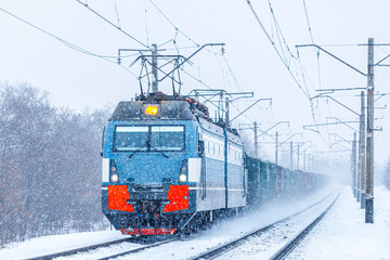 A gray locomotive with a long freight train is going to the railway station. Blizzard. Freight railway transportation of goods. Winter weather.