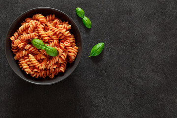 Fusilli pasta cooked with tomato sauce and basil in bowl, on dark background, top view, space to...