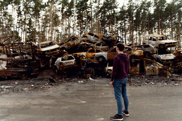 Ukrainian man stands against the background of broken and burnt cars affected by Russian missiles. dump of cars. the guy looks at the broken cars. bomb fragment holes. war in ukraine