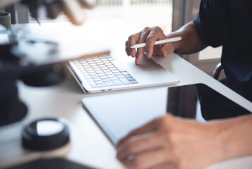 Scientist working on laptop computer and using digital tablet with microscope. Doctor or technician recording lab test on tablet pc in hospital laboratory