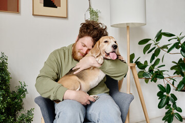 Young bearded man sitting on armchair in room with plants embracing his dog and relaxing