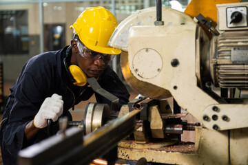 Professional African American engineer or worker wearing a hard hat and using machine for cutting the piece of metal pipe in the factory.