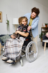 Fototapeta na wymiar Young bearded guy caring about his girlfriend with disability at home, she sitting in wheelchair and holding dog in her arms