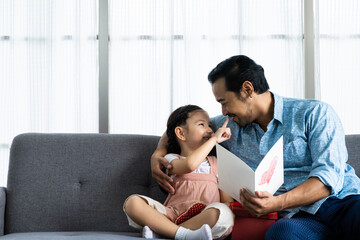 Child Asian girl give a present to his dad while smile, hug and kiss inside of the living room. Happy moment with male parent, family relationship. Father day