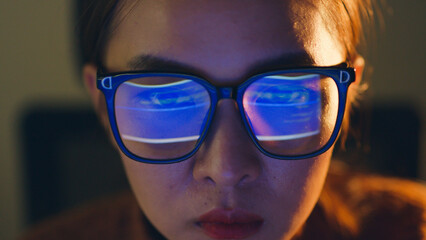 Close up of face of young Asian woman, developer programmer, software engineer, IT support, wearing glasses working hard at night overtime on computer to check coding in bugging system. - obrazy, fototapety, plakaty