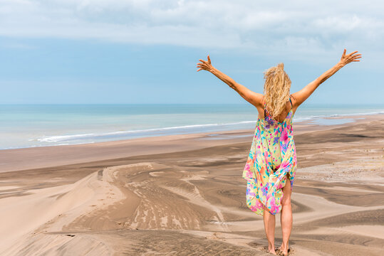 Photo with copy space of the back of a blonde woman raising arms on the top a dune facing the ocean