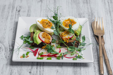 Bread toast, boiled eggs, avocado slice, pea microgreens and fresh radish on a white plate in a cafe, breakfast time