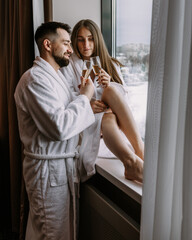 Couple wearing bathrobes in hotel standing near window with glasses of champagne