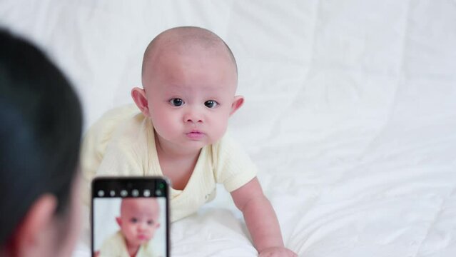 Authentic shot of Mother taking photo or video, cute Asian baby with mobile phone on white bed at home, happy smile face. Little innocent new infant adorable. Technology lifestyle parenthood concept.