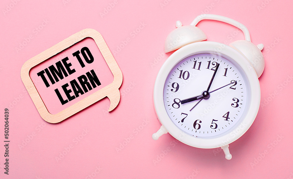Poster on a delicate pink background, a white alarm clock and a wooden frame with the text time to learn