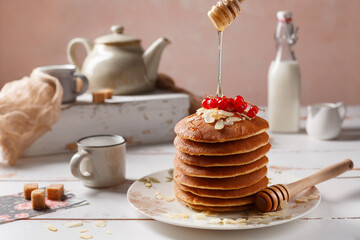 Miel qui coule sur les crêpes. Crêpes sur une assiette couverts de groseilles et amandes effilées.  Le bouteille de lait à coté et une tasse à thé sur une table rustique.