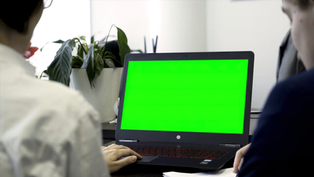 Young Women Working In Office, Sitting In Chairs In Front Of Computer With Green Screen, Rear View. Two Girls Co Workers Looking At Computer Monitor With Chroma Key Over White Wall Background.