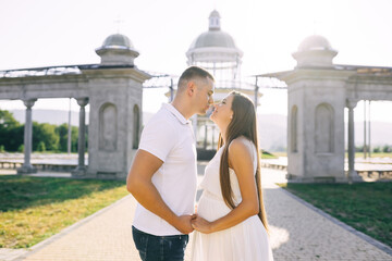 Young family of beautiful pregnant woman in white dress and man kissing while walking the streets of the city.