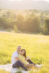 A happy couple a man with a pregnant woman sits in a meadow, hugging their stomachs, among the grass during a walk