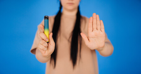 a girl on a blue background holds a disposable cigarette in one hand and shows a stop to smoking with the other hand. the concept of a healthy lifestyle