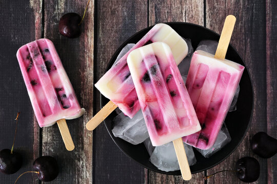 Plate of healthy cherry yogurt popsicles. Overhead view table scene over a rustic dark wood background.