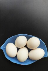 Heap or group of several isolated farm fresh organic white raw duck or hen eggs on a blue plate with black table background surface and copy space. Beautiful vertical closeup macro top view.