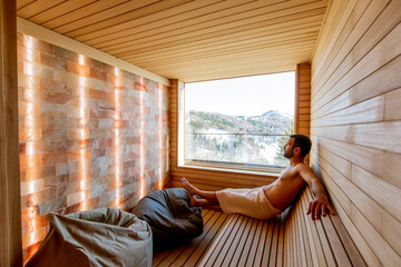 Young man relaxing in the sauna and watching winter forest through the window