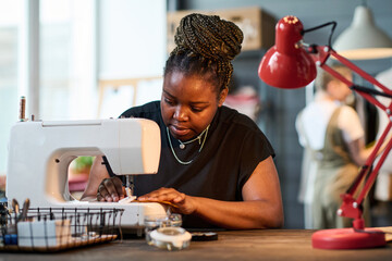 Young serious female tailor using electric sewing machine by her workplace against workshop interior and working colleague