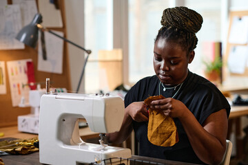 Contemporary serious female tailor with yellow fabric cutting part of new piece of clothing while sitting in front of electric sewing machine