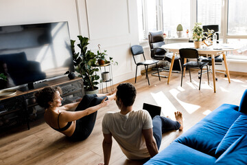 Back view couple stretching watching fitness video tutorial online on laptop. Fit man and woman doing yoga workout at home sitting on floor in living room together, hold hands.