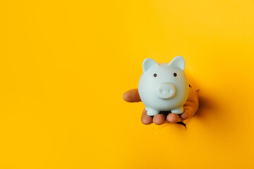Hand with a blue piggy bank through a yellow paper hole with copy space