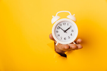 Hand holds a white alarm clock through a paper hole in yellow background with copy space