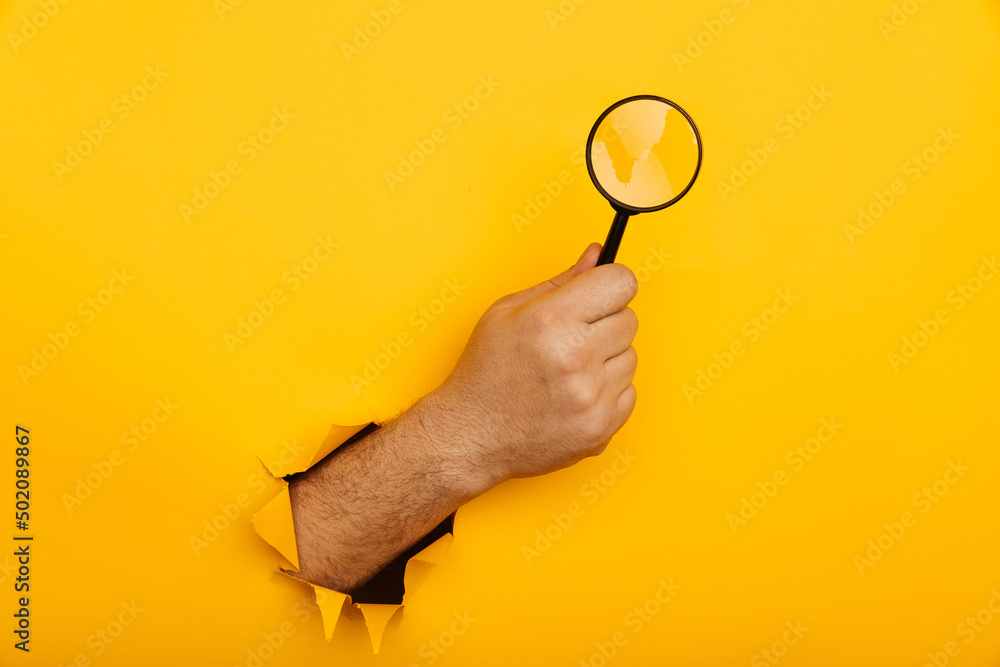 Wall mural male hand holds black magnifying glass in torn hole of yellow background. searching information conc