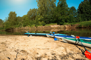 Obraz na płótnie Canvas Tourist canoes with paddles stand on the river coast in summer on a water hike. Rafting on inflatable and frame double and triple kayak boats, family trip, extreme adventure in summer
