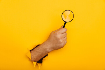 Male hand holds black magnifying glass in torn hole of yellow background. Searching information...