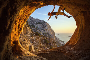 Young woman free solo climbing in cave with beautiful sea view in background - obrazy, fototapety, plakaty