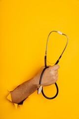 Hand holds a stethoscope out of a hole torn in yellow paper wall, vertical image