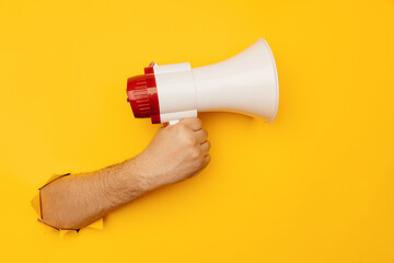 Male hand holds a megaphone from a hole in the wall on a yellow background. Advertising concept