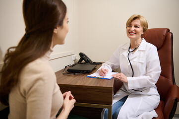 Fototapeta premium European experienced female physician consulting a young woman , filling out a medical form and prescribing medication at appointment in hospital