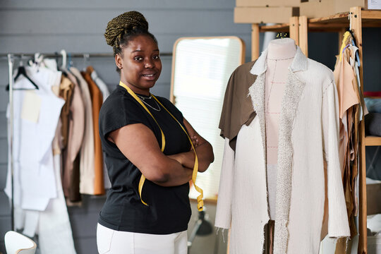 Young successful African American seamstress or tailor standng by mannequin with unfinished white coat on in workshop