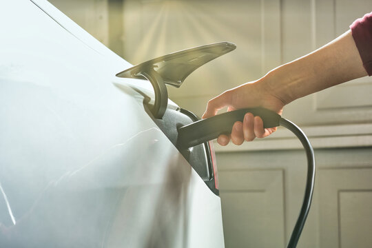 Man Plugging In Cable To Socket And Charging White Electric Car At Home In Garage Under Sunlight.  EV Green Energy Background. 