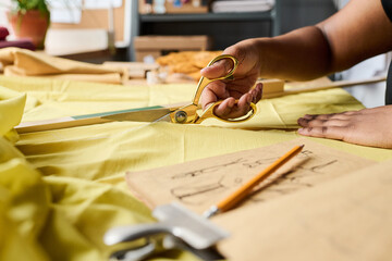 Hand of young black woman holding scissors while cutting yellow fabric on workplace during creation of new fashion collection